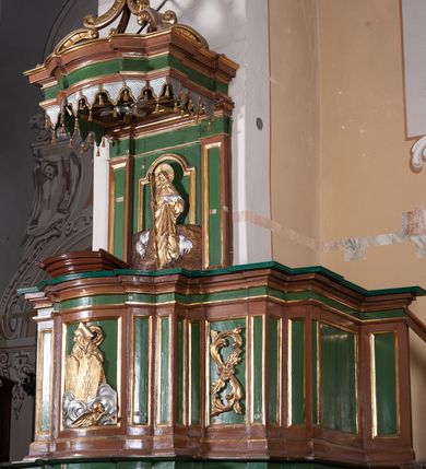 Zdjęcie nr 2: Ambona przyścienna składająca się z kosza, zaplecka, baldachimu i schodów z balustradą, zawieszona na lewej ścianie prezbiterium. Kosz na planie zbliżonym do prostokąta, o ściankach prostych i wklęsłych z uskokami w narożach, dekorowanymi płycinami z plakietami ornamentalnymi. Na największej płycinie przedstawienie Boga Ojca trzymającego tablice 10 przykazań, pośród obłoków, z trąbiącym aniołem w prawym dolnym narożniku. Kosz zamyka pełne belkowanie z silnie wysuniętym gzymsem. Podstawa kosza z łączących się pośrodku wolut, z pozłacaną szyszką u dołu. Zaplecek w kształcie prostokąta z prostokątną płyciną zamknięta łukiem nadwieszonym, wewnątrz której płaskorzeźbione przedstawienie Chrystusa Dobrego Pasterza. Pole flankują pilastry z płycinami. Baldachim powtarzający plan kosza, z podwieszonym lambrekinem i zwieńczony postumentem utworzonym ze stylizowanych wolut, na którym posadowiono figurę modlącego się anioła w długiej, srebrnej sukni. W podniebiu baldachimu znajduje się rzeźba srebrnej gołębicy Ducha Świętego na tle glorii promienistej. Struktura w kolorze brązowym, płyciny ciemnozielone, detal architektoniczny i ornamenty pozłacane i posrebrzane.
