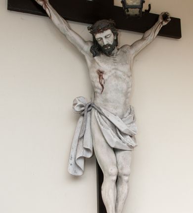 Zdjęcie nr 2: Na skrzyżowaniu ramion postać zmarłego Chrystusa, przybitego trzema gwoździami do krzyża. Ciało w delikatnym zwisie, o szeroko rozłożonych ramionach i rozciągniętej, prostej sylwetce. Głowa w koronie cierniowej opada na prawe ramię. Twarz o precyzyjnie oddanych rysach, okolona zarostem; włosy w postaci pukli opadają na plecy i prawe ramię. Ciało oddane z dbałością o detale anatomiczne, z zaznaczonymi śladami męki – krwią wypływającą z miejsc przebić i z rany na prawym boku. Białe perizonium w formie tkaniny opasującej biodra, drapowanej w spływające ku ziemi fałdy, z węzłem i zwisem na prawym boku. Titulus w kształcie banderoli z napisem „INRI”. Polichromia w partiach ciała naturalistyczna.