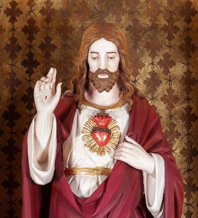 Zdjęcie nr 1: Rzeźba pełnoplastyczna przedstawiająca Chrystusa, stojącego centralnie, z lewą nogą lekko ugiętą w kolanie. Ręce uniesione, prawa w geście błogosławieństwa, ze znakiem męki, lewa odchyla płaszcz, by ukazać serce oplecione koroną cierniową, z płomieniem, zza którego widać krzyż i na tle glorii. Głowa lekko pochylona, oczy skierowane w dół, nos prosty, usta wąskie. Twarz okala krótki brązowy zarost oraz falowane włosy opadające na ramiona i plecy. Chrystus jest ubrany w białą tunikę z długimi rękawami, odsłaniającą nagie stopy. Na to ma narzucony czerwony płaszcz, który odkrywa jedynie tors, fragmenty rękawów i tuniki przy prawej nodze. Polichromia w partiach ciała naturalistyczna.
