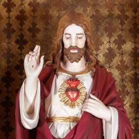 Zdjęcie nr 1: Rzeźba pełnoplastyczna przedstawiająca Chrystusa, stojącego centralnie, z lewą nogą lekko ugiętą w kolanie. Ręce uniesione, prawa w geście błogosławieństwa, ze znakiem męki, lewa odchyla płaszcz, by ukazać serce oplecione koroną cierniową, z płomieniem, zza którego widać krzyż i na tle glorii. Głowa lekko pochylona, oczy skierowane w dół, nos prosty, usta wąskie. Twarz okala krótki brązowy zarost oraz falowane włosy opadające na ramiona i plecy. Chrystus jest ubrany w białą tunikę z długimi rękawami, odsłaniającą nagie stopy. Na to ma narzucony czerwony płaszcz, który odkrywa jedynie tors, fragmenty rękawów i tuniki przy prawej nodze. Polichromia w partiach ciała naturalistyczna.