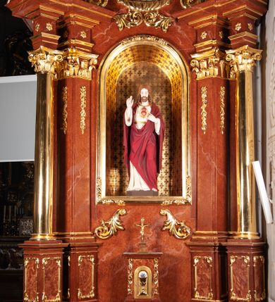 Zdjęcie nr 2: Rzeźba pełnoplastyczna przedstawiająca Chrystusa, stojącego centralnie, z lewą nogą lekko ugiętą w kolanie. Ręce uniesione, prawa w geście błogosławieństwa, ze znakiem męki, lewa odchyla płaszcz, by ukazać serce oplecione koroną cierniową, z płomieniem, zza którego widać krzyż i na tle glorii. Głowa lekko pochylona, oczy skierowane w dół, nos prosty, usta wąskie. Twarz okala krótki brązowy zarost oraz falowane włosy opadające na ramiona i plecy. Chrystus jest ubrany w białą tunikę z długimi rękawami, odsłaniającą nagie stopy. Na to ma narzucony czerwony płaszcz, który odkrywa jedynie tors, fragmenty rękawów i tuniki przy prawej nodze. Polichromia w partiach ciała naturalistyczna.