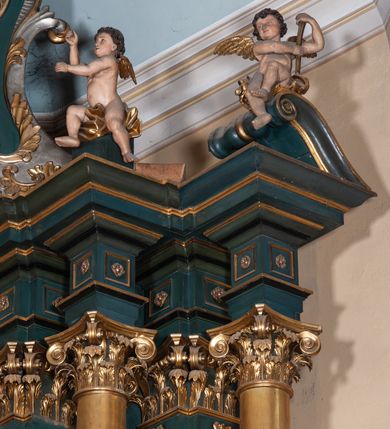 Zdjęcie nr 15: Ołtarz architektoniczny, jednoosiowy, jednokondygnacyjny ze zwieńczeniem, na podwójnym cokole. Mensa prostopadłościenna z antepedium dekorowanym płaskorzeźbionym przedstawieniem Jezusa rozdającego Komunię Świętą. Tabernakulum złożone z prostopadłościennej, obitej blachą skrzynki z drzwiczkami dekorowanymi literami „A” i „Ω” w górnej części i tablicami Dekalogu i otwartą księgą w dolnej, ujętymi winną latoroślą. Skrzynkę flankują prostopadłościany dekorowane na frontowych ściankach symbolami wiary i narzędziami męki, na których umieszczono klęczące, modlące się anioły. Między nimi ustawiono tron wystawienia w formie kolejnego prostopadłościanu, w którego frontowej ścianie znajduje się nisza wyłożona kratką z rozetami i ujęta dekoracją złożoną z księgi, tablic 10 przykazań i winnej latorośli.  Tron wystawienia zwieńczony gzymsem i postaciami dwóch aniołów, które klęczą i pochylają się w modlitewnych gestach. Oś nastawy flankują na przemian dwie pary pilastrów i dwie pary kolumn o kapitelach kompozytowych, ustawionych ukośnie względem lica ołtarza. Pomiędzy nimi znajdują się cztery figury (od lewej): św. Wojciecha, św. Jana Kantego, św. Kazimierza Jagiellończyka i św. Stanisława. W polu środkowym obraz z przedstawieniem świętych patronów Szymona i Judy Tadeusza w formie stojącego prostokąta zamkniętego łukiem odcinkowym w profilowanej, złoconej ramie ozdobionej rocaille’em. U spodu dwie ornamentalne ramki z motywem winnego grona z kluczami Piotrowymi po prawej i krzyżem ze stułą i lilią, po lewej. Kolumny dźwigają wyłamane belkowanie, przerwane w partii środkowej. Górną partię belkowania na osiach kolumn wieńczą putta posadowione na wolutach. Cokół dekorowany płycinami z rocaille’em, trzony pilastrów festonami utworzonymi z wstęgi i kiści winnego grona. W partii fryzu płyciny z rozetami. Fantazyjne zwieńczenie w formie zbliżonej do stojącego prostokąta. W nim, na obłoku półpostać błogosławiącego Boga Ojca w trójkątnym nimbie, z jabłkiem królewskim w formie globu zwieńczonego krzyżem w lewej dłoni. Poniżej gołębica Ducha Świętego w glorii promienistej. Kondygnacje od zwieńczenia oddziela kartusz utworzony z ornamentu rocaille’owego i wstęgi. Struktura polichromowana na kolor ciemnoniebieski, ze złoconą ornamentyką i złoconymi i srebrzonymi detalami.