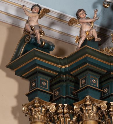 Zdjęcie nr 14: Ołtarz architektoniczny, jednoosiowy, jednokondygnacyjny ze zwieńczeniem, na podwójnym cokole. Mensa prostopadłościenna z antepedium dekorowanym płaskorzeźbionym przedstawieniem Jezusa rozdającego Komunię Świętą. Tabernakulum złożone z prostopadłościennej, obitej blachą skrzynki z drzwiczkami dekorowanymi literami „A” i „Ω” w górnej części i tablicami Dekalogu i otwartą księgą w dolnej, ujętymi winną latoroślą. Skrzynkę flankują prostopadłościany dekorowane na frontowych ściankach symbolami wiary i narzędziami męki, na których umieszczono klęczące, modlące się anioły. Między nimi ustawiono tron wystawienia w formie kolejnego prostopadłościanu, w którego frontowej ścianie znajduje się nisza wyłożona kratką z rozetami i ujęta dekoracją złożoną z księgi, tablic 10 przykazań i winnej latorośli.  Tron wystawienia zwieńczony gzymsem i postaciami dwóch aniołów, które klęczą i pochylają się w modlitewnych gestach. Oś nastawy flankują na przemian dwie pary pilastrów i dwie pary kolumn o kapitelach kompozytowych, ustawionych ukośnie względem lica ołtarza. Pomiędzy nimi znajdują się cztery figury (od lewej): św. Wojciecha, św. Jana Kantego, św. Kazimierza Jagiellończyka i św. Stanisława. W polu środkowym obraz z przedstawieniem świętych patronów Szymona i Judy Tadeusza w formie stojącego prostokąta zamkniętego łukiem odcinkowym w profilowanej, złoconej ramie ozdobionej rocaille’em. U spodu dwie ornamentalne ramki z motywem winnego grona z kluczami Piotrowymi po prawej i krzyżem ze stułą i lilią, po lewej. Kolumny dźwigają wyłamane belkowanie, przerwane w partii środkowej. Górną partię belkowania na osiach kolumn wieńczą putta posadowione na wolutach. Cokół dekorowany płycinami z rocaille’em, trzony pilastrów festonami utworzonymi z wstęgi i kiści winnego grona. W partii fryzu płyciny z rozetami. Fantazyjne zwieńczenie w formie zbliżonej do stojącego prostokąta. W nim, na obłoku półpostać błogosławiącego Boga Ojca w trójkątnym nimbie, z jabłkiem królewskim w formie globu zwieńczonego krzyżem w lewej dłoni. Poniżej gołębica Ducha Świętego w glorii promienistej. Kondygnacje od zwieńczenia oddziela kartusz utworzony z ornamentu rocaille’owego i wstęgi. Struktura polichromowana na kolor ciemnoniebieski, ze złoconą ornamentyką i złoconymi i srebrzonymi detalami.