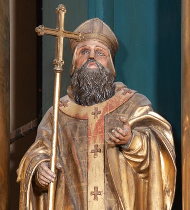 Zdjęcie nr 1: Figura ścięta z tyłu, na cokole. Święty Wojciech został ukazany w pozycji stojącej z uniesionymi rękami, w prawej trzyma krzyż misyjny. Twarz ma pociągłą, brwi wąskie, oczy szeroko otwarte, głęboko osadzone, nos prosty, wąski i długą, sięgającą dekoltu brodę. Jest ubrany w sutannę, komżę, ornat, stułę i paliusz; na głowie ma infułę. Polichromia w partiach ciała naturalistyczna, szaty i atrybuty złocone. 