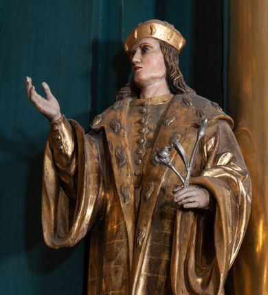 Zdjęcie nr 1: Rzeźba ścięta na cokole przedstawiająca stojącego frontalnie św. Kazimierza. Święty lewą stopę wystawia przed prawą, ma lekko uniesioną głowę, oczy skierowane w górę, nos prosty, usta wąskie. Jasnobrązowe włosy spływają mu falami na kark i ramiona, na głowie mitra książęca. Prawą rękę wyciąga przed siebie, w lewej, zgiętej w łokciu trzyma kwiat lilii. Jest ubrany w płaszcz zdobiony na kołnierzu motywami imitującymi cętki na futrze gronostajowym. Spod niego wystaje fragment zapinanej na guziki tuniki oraz wysokie buty. Polichromia w partiach ciała naturalistyczna, szaty złocone, lilia srebrzona. 