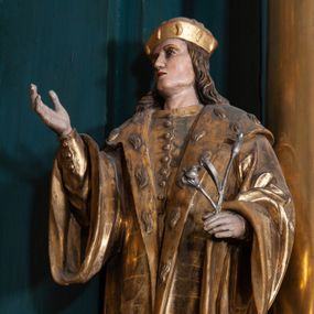Zdjęcie nr 1: Rzeźba ścięta na cokole przedstawiająca stojącego frontalnie św. Kazimierza. Święty lewą stopę wystawia przed prawą, ma lekko uniesioną głowę, oczy skierowane w górę, nos prosty, usta wąskie. Jasnobrązowe włosy spływają mu falami na kark i ramiona, na głowie mitra książęca. Prawą rękę wyciąga przed siebie, w lewej, zgiętej w łokciu trzyma kwiat lilii. Jest ubrany w płaszcz zdobiony na kołnierzu motywami imitującymi cętki na futrze gronostajowym. Spod niego wystaje fragment zapinanej na guziki tuniki oraz wysokie buty. Polichromia w partiach ciała naturalistyczna, szaty złocone, lilia srebrzona. 