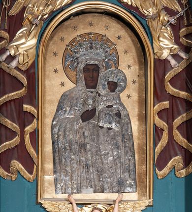Zdjęcie nr 2: Obraz w formie stojącego prostokąta zamkniętego łukiem pełnym z przedstawieniem Matki Boskiej w typie Częstochowskiej. Maria została ukazana frontalnie, w półpostaci. Patrzy na widza, twarz ma owalną, o wyrazistych rysach, ciemnej karnacji, długim nosie, ciemnych oczach i niewielkich ustach. Prawą ręka wskazuje na trzymane na lewej ręce Dzieciątko Jezus zwrócone w trzech czwartych w prawo, z głową odwróconą do widza. Twarz ma okrągłą, o ciemnej karnacji i wyrazistych rysach, na prawym policzku dwa nacięcia. Włosy brązowe, kręcone, zaczesane na twarz. Lewą rączką podtrzymuje księgę, prawą ma uniesioną w geście błogosławieństwa. Ubrania postaci oraz nimby i korony wykonane są ze srebrnej blachy, trybowanej w ornament roślinny, podkreślonej perełkowaniem na krawędziach i kameryzowanej w partii lamowań oraz koron. Maria ma pod szyją zawieszone sznury pereł. Nimb Dzieciątka jest wykonany z blachy, Marii złocony, oba promieniste. Obie postacie mają otwarte korony przytrzymywane przez niewielkie anioły; koronę Marii dekoruje dodatkowo pięć płaskorzeźbionych, złoconych gwiazd. Wokół głów obu postaci 12 gwiazd, tło jednolite – złote. 