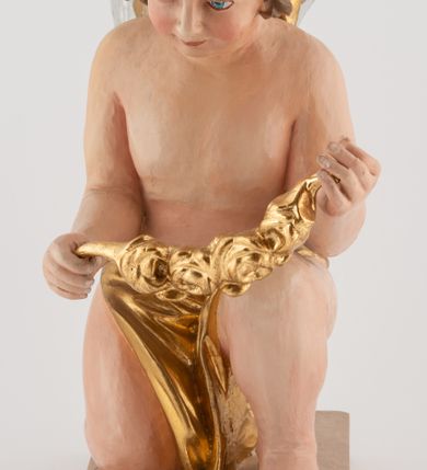 Zdjęcie nr 1: Pełnoplastyczna figura uskrzydlonego anioła ustawiona na niskim prostopadłościennym, beżowym cokole o ściętych narożach. Anioł zwrócony w prawo, klęczący na prawej nodze, z głową pochyloną delikatnie do przodu, z girlandą w dłoniach. Twarz okrągła o pełnych policzkach, z długim nosem i dużymi oczami. Włosy średniej długości, brązowe, zawinięte w duże loki do tyłu i z jednym kosmykiem na czole. Wokół bioder ma założoną złotą chustę. Polichromia w odsłoniętych partiach ciała naturalistyczna, końcówki skrzydeł srebrzone.