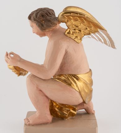 Zdjęcie nr 3: Pełnoplastyczna figura uskrzydlonego anioła ustawiona na niskim prostopadłościennym, beżowym cokole o ściętych narożach. Anioł zwrócony w prawo, klęczący na prawej nodze, z głową pochyloną delikatnie do przodu, z girlandą w dłoniach. Twarz okrągła o pełnych policzkach, z długim nosem i dużymi oczami. Włosy średniej długości, brązowe, zawinięte w duże loki do tyłu i z jednym kosmykiem na czole. Wokół bioder ma założoną złotą chustę. Polichromia w odsłoniętych partiach ciała naturalistyczna, końcówki skrzydeł srebrzone.