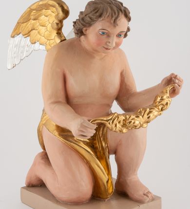 Zdjęcie nr 4: Pełnoplastyczna figura uskrzydlonego anioła ustawiona na niskim prostopadłościennym, beżowym cokole o ściętych narożach. Anioł zwrócony w prawo, klęczący na prawej nodze, z głową pochyloną delikatnie do przodu, z girlandą w dłoniach. Twarz okrągła o pełnych policzkach, z długim nosem i dużymi oczami. Włosy średniej długości, brązowe, zawinięte w duże loki do tyłu i z jednym kosmykiem na czole. Wokół bioder ma założoną złotą chustę. Polichromia w odsłoniętych partiach ciała naturalistyczna, końcówki skrzydeł srebrzone.