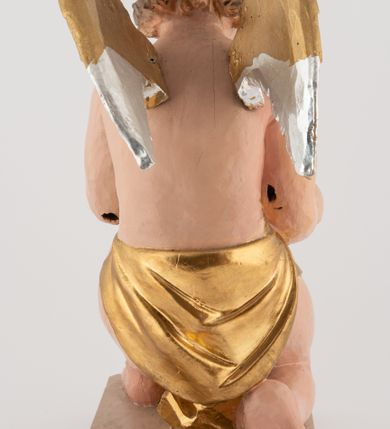 Zdjęcie nr 6: Pełnoplastyczna figura uskrzydlonego anioła ustawiona na niskim prostopadłościennym, beżowym cokole o ściętych narożach. Anioł zwrócony w prawo, klęczący na prawej nodze, z głową pochyloną delikatnie do przodu, z girlandą w dłoniach. Twarz okrągła o pełnych policzkach, z długim nosem i dużymi oczami. Włosy średniej długości, brązowe, zawinięte w duże loki do tyłu i z jednym kosmykiem na czole. Wokół bioder ma założoną złotą chustę. Polichromia w odsłoniętych partiach ciała naturalistyczna, końcówki skrzydeł srebrzone.