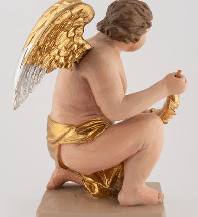 Zdjęcie nr 2: Pełnoplastyczna figura uskrzydlonego anioła ustawiona na niskim prostopadłościennym, beżowym cokole o ściętych narożach. Anioł zwrócony w lewo, klęczący na lewej nodze, z głową pochyloną delikatnie do przodu, z girlandą w dłoniach. Twarz okrągła o pełnych policzkach, z długim nosem i dużymi oczami. Włosy średniej długości, brązowe, zawinięte w duże loki do tyłu i z jednym kosmykiem na czole. Wokół bioder ma założoną złotą chustę. Polichromia w odsłoniętych partiach ciała naturalistyczna, końcówki skrzydeł srebrzone.