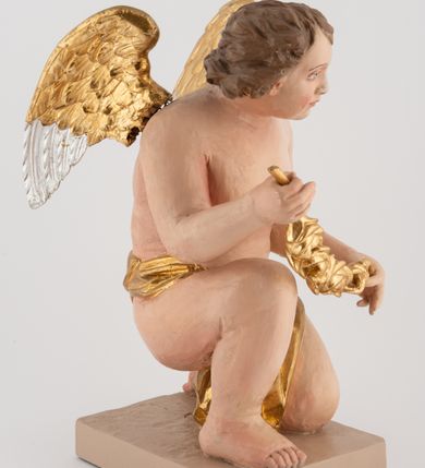 Zdjęcie nr 4: Pełnoplastyczna figura uskrzydlonego anioła ustawiona na niskim prostopadłościennym, beżowym cokole o ściętych narożach. Anioł zwrócony w lewo, klęczący na lewej nodze, z głową pochyloną delikatnie do przodu, z girlandą w dłoniach. Twarz okrągła o pełnych policzkach, z długim nosem i dużymi oczami. Włosy średniej długości, brązowe, zawinięte w duże loki do tyłu i z jednym kosmykiem na czole. Wokół bioder ma założoną złotą chustę. Polichromia w odsłoniętych partiach ciała naturalistyczna, końcówki skrzydeł srebrzone.