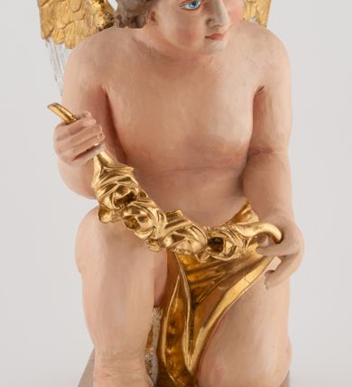 Zdjęcie nr 5: Pełnoplastyczna figura uskrzydlonego anioła ustawiona na niskim prostopadłościennym, beżowym cokole o ściętych narożach. Anioł zwrócony w lewo, klęczący na lewej nodze, z głową pochyloną delikatnie do przodu, z girlandą w dłoniach. Twarz okrągła o pełnych policzkach, z długim nosem i dużymi oczami. Włosy średniej długości, brązowe, zawinięte w duże loki do tyłu i z jednym kosmykiem na czole. Wokół bioder ma założoną złotą chustę. Polichromia w odsłoniętych partiach ciała naturalistyczna, końcówki skrzydeł srebrzone.