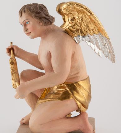 Zdjęcie nr 6: Pełnoplastyczna figura uskrzydlonego anioła ustawiona na niskim prostopadłościennym, beżowym cokole o ściętych narożach. Anioł zwrócony w lewo, klęczący na lewej nodze, z głową pochyloną delikatnie do przodu, z girlandą w dłoniach. Twarz okrągła o pełnych policzkach, z długim nosem i dużymi oczami. Włosy średniej długości, brązowe, zawinięte w duże loki do tyłu i z jednym kosmykiem na czole. Wokół bioder ma założoną złotą chustę. Polichromia w odsłoniętych partiach ciała naturalistyczna, końcówki skrzydeł srebrzone.