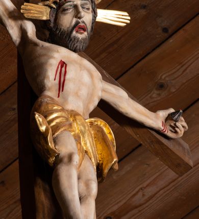 Zdjęcie nr 6: Pełnoplastyczna figura Chrystusa przybita do krzyża trzema gwoździami. Postać ukazana w łagodnym zwisie, ramiona ułożone w kształt litery „Y”, dłonie o lekko zaciśniętych palcach. Głowa, przechylona na prawą stronę, nogi skrzyżowane z prawą stopą założoną na lewą. Sylwetka ciała mocno wychudzona, z wydatną, wysuniętą do przodu klatką piersiową i wciętą talią. Twarz pociągła o wyrazistych rysach, okolona krótką brodą. Oczy o opadłych kącikach, usta rozchylone. Wokół bioder krótkie perizonium z fałdą przechodzącą między udami i zawiniętą z tyłu prawej nogi. Na głowie korona cierniowa, a wokół nimb krzyżowy. Karnacja ciała pokryta polichromią w naturalnym jasnokremowym odcieniu z zaznaczonymi śladami męki na dłoniach, stopach, prawym boku i czole. Perizonium, promienie w nimbie i korona cierniowa złote.