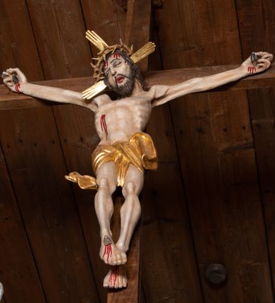 Zdjęcie nr 2: Pełnoplastyczna figura Chrystusa przybita do krzyża trzema gwoździami. Postać ukazana w łagodnym zwisie, ramiona ułożone w kształt litery „Y”, dłonie o lekko zaciśniętych palcach. Głowa, przechylona na prawą stronę, nogi skrzyżowane z prawą stopą założoną na lewą. Sylwetka ciała mocno wychudzona, z wydatną, wysuniętą do przodu klatką piersiową i wciętą talią. Twarz pociągła o wyrazistych rysach, okolona krótką brodą. Oczy o opadłych kącikach, usta rozchylone. Wokół bioder krótkie perizonium z fałdą przechodzącą między udami i zawiniętą z tyłu prawej nogi. Na głowie korona cierniowa, a wokół nimb krzyżowy. Karnacja ciała pokryta polichromią w naturalnym jasnokremowym odcieniu z zaznaczonymi śladami męki na dłoniach, stopach, prawym boku i czole. Perizonium, promienie w nimbie i korona cierniowa złote.