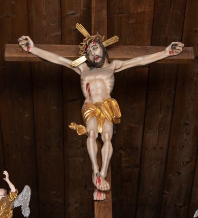 Zdjęcie nr 1: Pełnoplastyczna figura Chrystusa przybita do krzyża trzema gwoździami. Postać ukazana w łagodnym zwisie, ramiona ułożone w kształt litery „Y”, dłonie o lekko zaciśniętych palcach. Głowa, przechylona na prawą stronę, nogi skrzyżowane z prawą stopą założoną na lewą. Sylwetka ciała mocno wychudzona, z wydatną, wysuniętą do przodu klatką piersiową i wciętą talią. Twarz pociągła o wyrazistych rysach, okolona krótką brodą. Oczy o opadłych kącikach, usta rozchylone. Wokół bioder krótkie perizonium z fałdą przechodzącą między udami i zawiniętą z tyłu prawej nogi. Na głowie korona cierniowa, a wokół nimb krzyżowy. Karnacja ciała pokryta polichromią w naturalnym jasnokremowym odcieniu z zaznaczonymi śladami męki na dłoniach, stopach, prawym boku i czole. Perizonium, promienie w nimbie i korona cierniowa złote.