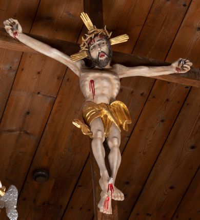 Zdjęcie nr 5: Pełnoplastyczna figura Chrystusa przybita do krzyża trzema gwoździami. Postać ukazana w łagodnym zwisie, ramiona ułożone w kształt litery „Y”, dłonie o lekko zaciśniętych palcach. Głowa, przechylona na prawą stronę, nogi skrzyżowane z prawą stopą założoną na lewą. Sylwetka ciała mocno wychudzona, z wydatną, wysuniętą do przodu klatką piersiową i wciętą talią. Twarz pociągła o wyrazistych rysach, okolona krótką brodą. Oczy o opadłych kącikach, usta rozchylone. Wokół bioder krótkie perizonium z fałdą przechodzącą między udami i zawiniętą z tyłu prawej nogi. Na głowie korona cierniowa, a wokół nimb krzyżowy. Karnacja ciała pokryta polichromią w naturalnym jasnokremowym odcieniu z zaznaczonymi śladami męki na dłoniach, stopach, prawym boku i czole. Perizonium, promienie w nimbie i korona cierniowa złote.