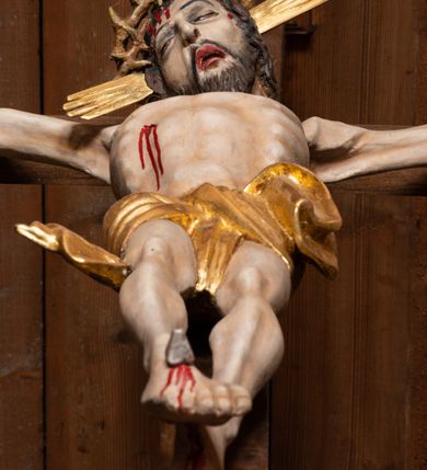 Zdjęcie nr 7: Pełnoplastyczna figura Chrystusa przybita do krzyża trzema gwoździami. Postać ukazana w łagodnym zwisie, ramiona ułożone w kształt litery „Y”, dłonie o lekko zaciśniętych palcach. Głowa, przechylona na prawą stronę, nogi skrzyżowane z prawą stopą założoną na lewą. Sylwetka ciała mocno wychudzona, z wydatną, wysuniętą do przodu klatką piersiową i wciętą talią. Twarz pociągła o wyrazistych rysach, okolona krótką brodą. Oczy o opadłych kącikach, usta rozchylone. Wokół bioder krótkie perizonium z fałdą przechodzącą między udami i zawiniętą z tyłu prawej nogi. Na głowie korona cierniowa, a wokół nimb krzyżowy. Karnacja ciała pokryta polichromią w naturalnym jasnokremowym odcieniu z zaznaczonymi śladami męki na dłoniach, stopach, prawym boku i czole. Perizonium, promienie w nimbie i korona cierniowa złote.
