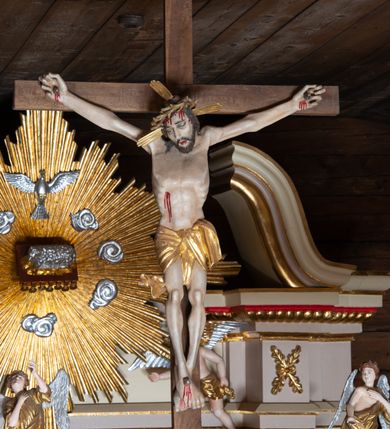 Zdjęcie nr 4: Pełnoplastyczna figura Chrystusa przybita do krzyża trzema gwoździami. Postać ukazana w łagodnym zwisie, ramiona ułożone w kształt litery „Y”, dłonie o lekko zaciśniętych palcach. Głowa, przechylona na prawą stronę, nogi skrzyżowane z prawą stopą założoną na lewą. Sylwetka ciała mocno wychudzona, z wydatną, wysuniętą do przodu klatką piersiową i wciętą talią. Twarz pociągła o wyrazistych rysach, okolona krótką brodą. Oczy o opadłych kącikach, usta rozchylone. Wokół bioder krótkie perizonium z fałdą przechodzącą między udami i zawiniętą z tyłu prawej nogi. Na głowie korona cierniowa, a wokół nimb krzyżowy. Karnacja ciała pokryta polichromią w naturalnym jasnokremowym odcieniu z zaznaczonymi śladami męki na dłoniach, stopach, prawym boku i czole. Perizonium, promienie w nimbie i korona cierniowa złote.