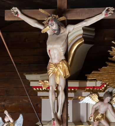 Zdjęcie nr 3: Pełnoplastyczna figura Chrystusa przybita do krzyża trzema gwoździami. Postać ukazana w łagodnym zwisie, ramiona ułożone w kształt litery „Y”, dłonie o lekko zaciśniętych palcach. Głowa, przechylona na prawą stronę, nogi skrzyżowane z prawą stopą założoną na lewą. Sylwetka ciała mocno wychudzona, z wydatną, wysuniętą do przodu klatką piersiową i wciętą talią. Twarz pociągła o wyrazistych rysach, okolona krótką brodą. Oczy o opadłych kącikach, usta rozchylone. Wokół bioder krótkie perizonium z fałdą przechodzącą między udami i zawiniętą z tyłu prawej nogi. Na głowie korona cierniowa, a wokół nimb krzyżowy. Karnacja ciała pokryta polichromią w naturalnym jasnokremowym odcieniu z zaznaczonymi śladami męki na dłoniach, stopach, prawym boku i czole. Perizonium, promienie w nimbie i korona cierniowa złote.