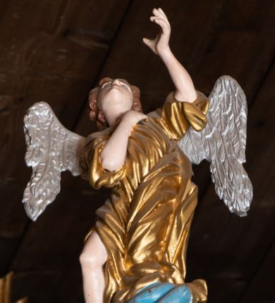 Zdjęcie nr 1: Pełnoplastyczna figura uskrzydlonego anioła ustawiona na niskim prostopadłościennym cokole. Anioł ukazany frontalnie na niebieskiej chmurze o silnie, esowato wygiętej sylwetce z głową uniesioną do góry, z prawą ręką złożoną na piersi, lewą ugiętą w łokciu uniesioną do góry. Twarz owalna o pełnych policzkach z drobnymi ustami i wyraźnie zaznaczonym podbródkiem. Włosy średniej długości, bujne, brązowe, zasłaniające uszy. Ubrany jest w złoconą tunikę z rękawami podwiniętymi do wysokości łokci, odsłaniającą prawą nogę. Polichromia w odsłoniętych partiach ciała naturalistyczna, skrzydła  srebrzone, suknia złocona, cokół i chmura malowane na niebiesko.