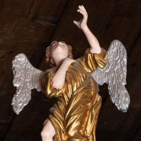 Zdjęcie nr 1: Pełnoplastyczna figura uskrzydlonego anioła ustawiona na niskim prostopadłościennym cokole. Anioł ukazany frontalnie na niebieskiej chmurze o silnie, esowato wygiętej sylwetce z głową uniesioną do góry, z prawą ręką złożoną na piersi, lewą ugiętą w łokciu uniesioną do góry. Twarz owalna o pełnych policzkach z drobnymi ustami i wyraźnie zaznaczonym podbródkiem. Włosy średniej długości, bujne, brązowe, zasłaniające uszy. Ubrany jest w złoconą tunikę z rękawami podwiniętymi do wysokości łokci, odsłaniającą prawą nogę. Polichromia w odsłoniętych partiach ciała naturalistyczna, skrzydła  srebrzone, suknia złocona, cokół i chmura malowane na niebiesko.