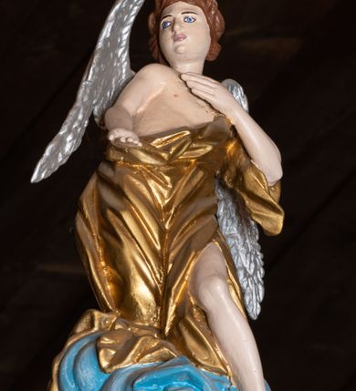 Zdjęcie nr 1: Pełnoplastyczna figura uskrzydlonego anioła ustawiona na niskim prostopadłościennym cokole. Anioł ukazany frontalnie na niebieskiej chmurze o silnie, esowato wygiętej sylwetce z głową zwróconą w prawą stronę, z prawą ręką opuszczoną wzdłuż ciała, a lewą ugiętą w łokciu i złożoną na piersi. Twarz szeroka o pełnych policzkach z drobnymi ustami i wyraźnie zaznaczonym podbródkiem. Włosy średniej długości, bujne, brązowe, zasłaniające uszy. Ubrany jest w złoconą tunikę, odsłaniającą prawe ramię, lewą nogę i tors. Polichromia w odsłoniętych partiach ciała naturalistyczna, skrzydła  srebrzone, suknia złocona, cokół i chmura malowane na niebiesko.