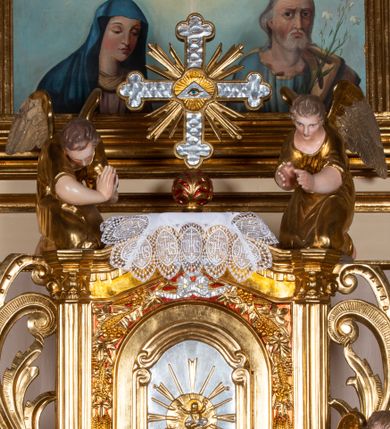 Zdjęcie nr 1: Pełnoplastyczne figury dwóch uskrzydlonych aniołów. Anioły zwrócone ku tabernakulum, ukazane w postawie klęczącej, z głową pochyloną delikatnie do przodu, z rękami ugiętymi w łokciach i wysuniętymi do przodu, które składają w geście modlitwy. Twarze podłużne o pełnych policzkach, z długimi nosami i dużymi niebieskimi oczami. Włosy krótkie, brązowe, falowane, zasłaniające uszy. Ubrane są w długie suknie z rękawami podwiniętymi do wysokości łokcia, przewiązane w pasie sznurami. Polichromia w odsłoniętych partiach ciała naturalistyczna, końcówki skrzydeł srebrzone, suknia złocona.
