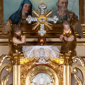 Zdjęcie nr 1: Pełnoplastyczne figury dwóch uskrzydlonych aniołów. Anioły zwrócone ku tabernakulum, ukazane w postawie klęczącej, z głową pochyloną delikatnie do przodu, z rękami ugiętymi w łokciach i wysuniętymi do przodu, które składają w geście modlitwy. Twarze podłużne o pełnych policzkach, z długimi nosami i dużymi niebieskimi oczami. Włosy krótkie, brązowe, falowane, zasłaniające uszy. Ubrane są w długie suknie z rękawami podwiniętymi do wysokości łokcia, przewiązane w pasie sznurami. Polichromia w odsłoniętych partiach ciała naturalistyczna, końcówki skrzydeł srebrzone, suknia złocona.