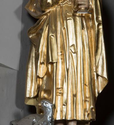 Zdjęcie nr 1: Figura św. Jana znajduje się ponad bramką prowadzącą za ołtarz, na postumencie ozdobionym od frontu ornamentem małżowinowym. Ewangelista jest ukazany w całej postaci, w pozycji stojącej, lekko przechylony w swoją lewą stronę, z orłem siedzącym u jego stóp. Święty w lewej dłoni trzyma kielich, z którego czary wyłania się wąż. Prawa ręka jest uniesiona w geście błogosławieństwa. Twarz świętego jest owalna, z wysokim czołem, prostym nosem oraz głęboko osadzonymi oczami. Jego usta są wąskie i zaciśnięte. Włosy opadają na ramiona w postaci pofalowanych pukli. Ewangelista nie ma obuwia, odziany jest w przewiązaną w pasie tunikę sięgającą kostek z kołnierzem i wąskimi rękawami. Na tunikę założony jest płaszcz, przerzucony przez lewe ramię. W opracowaniu tuniki dominują pionowe, równoległe, rurkowate fałdy, płaszcz kształtowany jest poprzez załamane fałdy. Karnacja ciała świętego kremowa, włosy brązowe, szaty złote. 