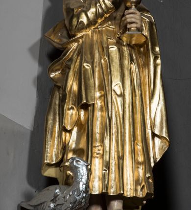 Zdjęcie nr 2: Figura św. Jana znajduje się ponad bramką prowadzącą za ołtarz, na postumencie ozdobionym od frontu ornamentem małżowinowym. Ewangelista jest ukazany w całej postaci, w pozycji stojącej, lekko przechylony w swoją lewą stronę, z orłem siedzącym u jego stóp. Święty w lewej dłoni trzyma kielich, z którego czary wyłania się wąż. Prawa ręka jest uniesiona w geście błogosławieństwa. Twarz świętego jest owalna, z wysokim czołem, prostym nosem oraz głęboko osadzonymi oczami. Jego usta są wąskie i zaciśnięte. Włosy opadają na ramiona w postaci pofalowanych pukli. Ewangelista nie ma obuwia, odziany jest w przewiązaną w pasie tunikę sięgającą kostek z kołnierzem i wąskimi rękawami. Na tunikę założony jest płaszcz, przerzucony przez lewe ramię. W opracowaniu tuniki dominują pionowe, równoległe, rurkowate fałdy, płaszcz kształtowany jest poprzez załamane fałdy. Karnacja ciała świętego kremowa, włosy brązowe, szaty złote. 