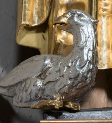 Zdjęcie nr 5: Figura św. Jana znajduje się ponad bramką prowadzącą za ołtarz, na postumencie ozdobionym od frontu ornamentem małżowinowym. Ewangelista jest ukazany w całej postaci, w pozycji stojącej, lekko przechylony w swoją lewą stronę, z orłem siedzącym u jego stóp. Święty w lewej dłoni trzyma kielich, z którego czary wyłania się wąż. Prawa ręka jest uniesiona w geście błogosławieństwa. Twarz świętego jest owalna, z wysokim czołem, prostym nosem oraz głęboko osadzonymi oczami. Jego usta są wąskie i zaciśnięte. Włosy opadają na ramiona w postaci pofalowanych pukli. Ewangelista nie ma obuwia, odziany jest w przewiązaną w pasie tunikę sięgającą kostek z kołnierzem i wąskimi rękawami. Na tunikę założony jest płaszcz, przerzucony przez lewe ramię. W opracowaniu tuniki dominują pionowe, równoległe, rurkowate fałdy, płaszcz kształtowany jest poprzez załamane fałdy. Karnacja ciała świętego kremowa, włosy brązowe, szaty złote. 