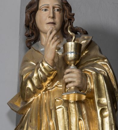 Zdjęcie nr 3: Figura św. Jana znajduje się ponad bramką prowadzącą za ołtarz, na postumencie ozdobionym od frontu ornamentem małżowinowym. Ewangelista jest ukazany w całej postaci, w pozycji stojącej, lekko przechylony w swoją lewą stronę, z orłem siedzącym u jego stóp. Święty w lewej dłoni trzyma kielich, z którego czary wyłania się wąż. Prawa ręka jest uniesiona w geście błogosławieństwa. Twarz świętego jest owalna, z wysokim czołem, prostym nosem oraz głęboko osadzonymi oczami. Jego usta są wąskie i zaciśnięte. Włosy opadają na ramiona w postaci pofalowanych pukli. Ewangelista nie ma obuwia, odziany jest w przewiązaną w pasie tunikę sięgającą kostek z kołnierzem i wąskimi rękawami. Na tunikę założony jest płaszcz, przerzucony przez lewe ramię. W opracowaniu tuniki dominują pionowe, równoległe, rurkowate fałdy, płaszcz kształtowany jest poprzez załamane fałdy. Karnacja ciała świętego kremowa, włosy brązowe, szaty złote. 