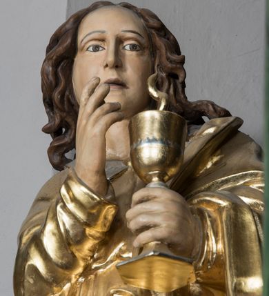 Zdjęcie nr 4: Figura św. Jana znajduje się ponad bramką prowadzącą za ołtarz, na postumencie ozdobionym od frontu ornamentem małżowinowym. Ewangelista jest ukazany w całej postaci, w pozycji stojącej, lekko przechylony w swoją lewą stronę, z orłem siedzącym u jego stóp. Święty w lewej dłoni trzyma kielich, z którego czary wyłania się wąż. Prawa ręka jest uniesiona w geście błogosławieństwa. Twarz świętego jest owalna, z wysokim czołem, prostym nosem oraz głęboko osadzonymi oczami. Jego usta są wąskie i zaciśnięte. Włosy opadają na ramiona w postaci pofalowanych pukli. Ewangelista nie ma obuwia, odziany jest w przewiązaną w pasie tunikę sięgającą kostek z kołnierzem i wąskimi rękawami. Na tunikę założony jest płaszcz, przerzucony przez lewe ramię. W opracowaniu tuniki dominują pionowe, równoległe, rurkowate fałdy, płaszcz kształtowany jest poprzez załamane fałdy. Karnacja ciała świętego kremowa, włosy brązowe, szaty złote. 