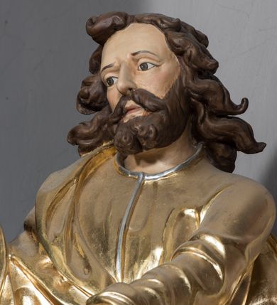 Zdjęcie nr 3: Figura św. Łukasza jest umieszczona ponad bramką prowadzącą za ołtarz, na postumencie, ozdobionym od frontu ornamentem małżowinowym. Święty ukazany jest w pozycji stojącej, lekko pochylony w przód, z prawą nogą ugiętą w kolanie i wspartą na leżącym u jego stóp wole. W prawej lekko zgiętej, wyciągniętej do przodu ręce trzyma ptasie pióro, lewą natomiast podtrzymuje księgę, która jest rozłożona i wsparta na kolanie. Twarz ewangelisty jest pociągła, z prostym nosem, szeroko otwartymi, dużymi oczami. Ponad wysokim czołem zaczepione są pofalowane pukle włosów uformowane w taki sposób, jak gdyby były rozwiane. Święty stoi boso, ma na sobie długą tunikę z dekoltem ozdobionym delikatną bordiurą oraz wąskimi rękawami, przewiązaną w pasie. Na wierzchu narzucony jest płaszcz spływający z prawego ramienia. Pod jego lewą ręką przywiązany do płaszcza kałamarz. W opracowaniu tuniki dominują pionowe, równoległe, rurkowate fałdy, a w przypadku płaszcza fałdy załamane i wygięte zgodnie z duktem tkaniny. Fragmenty ciała polichromowane naturalistycznie, szaty złocone, lamówki szat, wół i kałamarz srebrzone. 