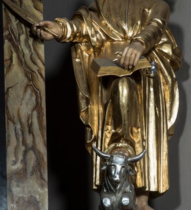 Zdjęcie nr 2: Figura św. Łukasza jest umieszczona ponad bramką prowadzącą za ołtarz, na postumencie, ozdobionym od frontu ornamentem małżowinowym. Święty ukazany jest w pozycji stojącej, lekko pochylony w przód, z prawą nogą ugiętą w kolanie i wspartą na leżącym u jego stóp wole. W prawej lekko zgiętej, wyciągniętej do przodu ręce trzyma ptasie pióro, lewą natomiast podtrzymuje księgę, która jest rozłożona i wsparta na kolanie. Twarz ewangelisty jest pociągła, z prostym nosem, szeroko otwartymi, dużymi oczami. Ponad wysokim czołem zaczepione są pofalowane pukle włosów uformowane w taki sposób, jak gdyby były rozwiane. Święty stoi boso, ma na sobie długą tunikę z dekoltem ozdobionym delikatną bordiurą oraz wąskimi rękawami, przewiązaną w pasie. Na wierzchu narzucony jest płaszcz spływający z prawego ramienia. Pod jego lewą ręką przywiązany do płaszcza kałamarz. W opracowaniu tuniki dominują pionowe, równoległe, rurkowate fałdy, a w przypadku płaszcza fałdy załamane i wygięte zgodnie z duktem tkaniny. Fragmenty ciała polichromowane naturalistycznie, szaty złocone, lamówki szat, wół i kałamarz srebrzone. 