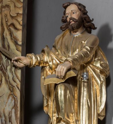 Zdjęcie nr 1: Figura św. Łukasza jest umieszczona ponad bramką prowadzącą za ołtarz, na postumencie, ozdobionym od frontu ornamentem małżowinowym. Święty ukazany jest w pozycji stojącej, lekko pochylony w przód, z prawą nogą ugiętą w kolanie i wspartą na leżącym u jego stóp wole. W prawej lekko zgiętej, wyciągniętej do przodu ręce trzyma ptasie pióro, lewą natomiast podtrzymuje księgę, która jest rozłożona i wsparta na kolanie. Twarz ewangelisty jest pociągła, z prostym nosem, szeroko otwartymi, dużymi oczami. Ponad wysokim czołem zaczepione są pofalowane pukle włosów uformowane w taki sposób, jak gdyby były rozwiane. Święty stoi boso, ma na sobie długą tunikę z dekoltem ozdobionym delikatną bordiurą oraz wąskimi rękawami, przewiązaną w pasie. Na wierzchu narzucony jest płaszcz spływający z prawego ramienia. Pod jego lewą ręką przywiązany do płaszcza kałamarz. W opracowaniu tuniki dominują pionowe, równoległe, rurkowate fałdy, a w przypadku płaszcza fałdy załamane i wygięte zgodnie z duktem tkaniny. Fragmenty ciała polichromowane naturalistycznie, szaty złocone, lamówki szat, wół i kałamarz srebrzone. 