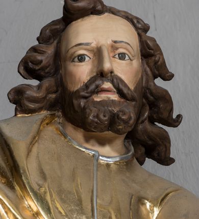 Zdjęcie nr 4: Figura św. Łukasza jest umieszczona ponad bramką prowadzącą za ołtarz, na postumencie, ozdobionym od frontu ornamentem małżowinowym. Święty ukazany jest w pozycji stojącej, lekko pochylony w przód, z prawą nogą ugiętą w kolanie i wspartą na leżącym u jego stóp wole. W prawej lekko zgiętej, wyciągniętej do przodu ręce trzyma ptasie pióro, lewą natomiast podtrzymuje księgę, która jest rozłożona i wsparta na kolanie. Twarz ewangelisty jest pociągła, z prostym nosem, szeroko otwartymi, dużymi oczami. Ponad wysokim czołem zaczepione są pofalowane pukle włosów uformowane w taki sposób, jak gdyby były rozwiane. Święty stoi boso, ma na sobie długą tunikę z dekoltem ozdobionym delikatną bordiurą oraz wąskimi rękawami, przewiązaną w pasie. Na wierzchu narzucony jest płaszcz spływający z prawego ramienia. Pod jego lewą ręką przywiązany do płaszcza kałamarz. W opracowaniu tuniki dominują pionowe, równoległe, rurkowate fałdy, a w przypadku płaszcza fałdy załamane i wygięte zgodnie z duktem tkaniny. Fragmenty ciała polichromowane naturalistycznie, szaty złocone, lamówki szat, wół i kałamarz srebrzone. 