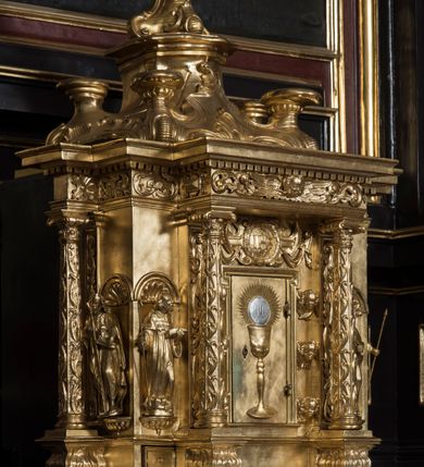 Zdjęcie nr 4: Tabernakulum w formie sześcianu, na niskim cokole, z dwukolumnowym portykiem utworzonym z pilastrów toskańskich i kolumn kompozytowych od frontu i dwóch analogicznych kolumn na tle pilastrów po bokach podtrzymujących przełamujące się na nimi belkowanie. Kolumny osadzone na niskich piedestałach, lekko wybrzuszonych, otoczonych liśćmi akantu. Kolumny pokryte symetrycznie układającymi i przeplatającymi się wokół pionowego trzpienia liśćmi i zwieńczone jońskimi kapitelami. Fryz belkowania zdobiony uskrzydloną główką aniołka, liściastymi wolutami, rautami w drobnych kartuszach i kostkowaniem. Drzwi tabernakulum zdobione są płaskorzeźbionym kielichem z hostią. Po bokach wypukło rzeźbione główki anielskie, a nad nimi okrągły medalion z literami IHS obwiedziony wieńcem laurowym, z elementami przechodzących między sobą listw kartuszowych i podwieszonych pod nie festonów. W ściankach naroży tabernakulum cztery nisze konchowe z muszlami wypełnione figurkami świętych. Jeden w wielbłądziej skórze trzymający księgę z barankiem przedstawia św. Jana Chrzciciela. Trzej pozostali są trudni do identyfikacji. Jeden trzyma otwartą księgę, a u jego stóp widnieje mitra książęca, drugi trzyma pastorał a u jego stóp leży infuła, trzeci z laską i kapeluszem u stóp (św. Jakub?). 


