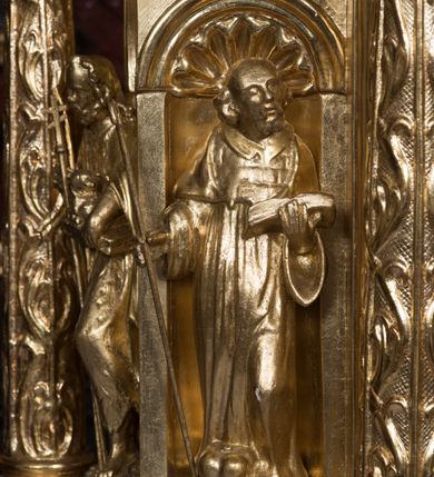 Zdjęcie nr 7: Tabernakulum w formie sześcianu, na niskim cokole, z dwukolumnowym portykiem utworzonym z pilastrów toskańskich i kolumn kompozytowych od frontu i dwóch analogicznych kolumn na tle pilastrów po bokach podtrzymujących przełamujące się na nimi belkowanie. Kolumny osadzone na niskich piedestałach, lekko wybrzuszonych, otoczonych liśćmi akantu. Kolumny pokryte symetrycznie układającymi i przeplatającymi się wokół pionowego trzpienia liśćmi i zwieńczone jońskimi kapitelami. Fryz belkowania zdobiony uskrzydloną główką aniołka, liściastymi wolutami, rautami w drobnych kartuszach i kostkowaniem. Drzwi tabernakulum zdobione są płaskorzeźbionym kielichem z hostią. Po bokach wypukło rzeźbione główki anielskie, a nad nimi okrągły medalion z literami IHS obwiedziony wieńcem laurowym, z elementami przechodzących między sobą listw kartuszowych i podwieszonych pod nie festonów. W ściankach naroży tabernakulum cztery nisze konchowe z muszlami wypełnione figurkami świętych. Jeden w wielbłądziej skórze trzymający księgę z barankiem przedstawia św. Jana Chrzciciela. Trzej pozostali są trudni do identyfikacji. Jeden trzyma otwartą księgę, a u jego stóp widnieje mitra książęca, drugi trzyma pastorał a u jego stóp leży infuła, trzeci z laską i kapeluszem u stóp (św. Jakub?). 


