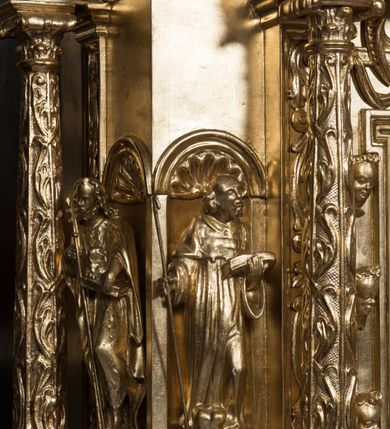 Zdjęcie nr 5: Tabernakulum w formie sześcianu, na niskim cokole, z dwukolumnowym portykiem utworzonym z pilastrów toskańskich i kolumn kompozytowych od frontu i dwóch analogicznych kolumn na tle pilastrów po bokach podtrzymujących przełamujące się na nimi belkowanie. Kolumny osadzone na niskich piedestałach, lekko wybrzuszonych, otoczonych liśćmi akantu. Kolumny pokryte symetrycznie układającymi i przeplatającymi się wokół pionowego trzpienia liśćmi i zwieńczone jońskimi kapitelami. Fryz belkowania zdobiony uskrzydloną główką aniołka, liściastymi wolutami, rautami w drobnych kartuszach i kostkowaniem. Drzwi tabernakulum zdobione są płaskorzeźbionym kielichem z hostią. Po bokach wypukło rzeźbione główki anielskie, a nad nimi okrągły medalion z literami IHS obwiedziony wieńcem laurowym, z elementami przechodzących między sobą listw kartuszowych i podwieszonych pod nie festonów. W ściankach naroży tabernakulum cztery nisze konchowe z muszlami wypełnione figurkami świętych. Jeden w wielbłądziej skórze trzymający księgę z barankiem przedstawia św. Jana Chrzciciela. Trzej pozostali są trudni do identyfikacji. Jeden trzyma otwartą księgę, a u jego stóp widnieje mitra książęca, drugi trzyma pastorał a u jego stóp leży infuła, trzeci z laską i kapeluszem u stóp (św. Jakub?). 


