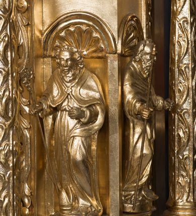 Zdjęcie nr 9: Tabernakulum w formie sześcianu, na niskim cokole, z dwukolumnowym portykiem utworzonym z pilastrów toskańskich i kolumn kompozytowych od frontu i dwóch analogicznych kolumn na tle pilastrów po bokach podtrzymujących przełamujące się na nimi belkowanie. Kolumny osadzone na niskich piedestałach, lekko wybrzuszonych, otoczonych liśćmi akantu. Kolumny pokryte symetrycznie układającymi i przeplatającymi się wokół pionowego trzpienia liśćmi i zwieńczone jońskimi kapitelami. Fryz belkowania zdobiony uskrzydloną główką aniołka, liściastymi wolutami, rautami w drobnych kartuszach i kostkowaniem. Drzwi tabernakulum zdobione są płaskorzeźbionym kielichem z hostią. Po bokach wypukło rzeźbione główki anielskie, a nad nimi okrągły medalion z literami IHS obwiedziony wieńcem laurowym, z elementami przechodzących między sobą listw kartuszowych i podwieszonych pod nie festonów. W ściankach naroży tabernakulum cztery nisze konchowe z muszlami wypełnione figurkami świętych. Jeden w wielbłądziej skórze trzymający księgę z barankiem przedstawia św. Jana Chrzciciela. Trzej pozostali są trudni do identyfikacji. Jeden trzyma otwartą księgę, a u jego stóp widnieje mitra książęca, drugi trzyma pastorał a u jego stóp leży infuła, trzeci z laską i kapeluszem u stóp (św. Jakub?). 


