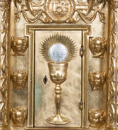 Zdjęcie nr 10: Tabernakulum w formie sześcianu, na niskim cokole, z dwukolumnowym portykiem utworzonym z pilastrów toskańskich i kolumn kompozytowych od frontu i dwóch analogicznych kolumn na tle pilastrów po bokach podtrzymujących przełamujące się na nimi belkowanie. Kolumny osadzone na niskich piedestałach, lekko wybrzuszonych, otoczonych liśćmi akantu. Kolumny pokryte symetrycznie układającymi i przeplatającymi się wokół pionowego trzpienia liśćmi i zwieńczone jońskimi kapitelami. Fryz belkowania zdobiony uskrzydloną główką aniołka, liściastymi wolutami, rautami w drobnych kartuszach i kostkowaniem. Drzwi tabernakulum zdobione są płaskorzeźbionym kielichem z hostią. Po bokach wypukło rzeźbione główki anielskie, a nad nimi okrągły medalion z literami IHS obwiedziony wieńcem laurowym, z elementami przechodzących między sobą listw kartuszowych i podwieszonych pod nie festonów. W ściankach naroży tabernakulum cztery nisze konchowe z muszlami wypełnione figurkami świętych. Jeden w wielbłądziej skórze trzymający księgę z barankiem przedstawia św. Jana Chrzciciela. Trzej pozostali są trudni do identyfikacji. Jeden trzyma otwartą księgę, a u jego stóp widnieje mitra książęca, drugi trzyma pastorał a u jego stóp leży infuła, trzeci z laską i kapeluszem u stóp (św. Jakub?). 


