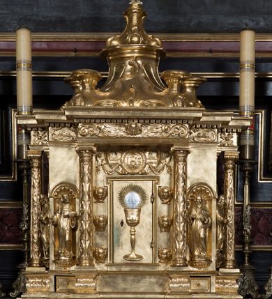 Zdjęcie nr 1: Tabernakulum w formie sześcianu, na niskim cokole, z dwukolumnowym portykiem utworzonym z pilastrów toskańskich i kolumn kompozytowych od frontu i dwóch analogicznych kolumn na tle pilastrów po bokach podtrzymujących przełamujące się na nimi belkowanie. Kolumny osadzone na niskich piedestałach, lekko wybrzuszonych, otoczonych liśćmi akantu. Kolumny pokryte symetrycznie układającymi i przeplatającymi się wokół pionowego trzpienia liśćmi i zwieńczone jońskimi kapitelami. Fryz belkowania zdobiony uskrzydloną główką aniołka, liściastymi wolutami, rautami w drobnych kartuszach i kostkowaniem. Drzwi tabernakulum zdobione są płaskorzeźbionym kielichem z hostią. Po bokach wypukło rzeźbione główki anielskie, a nad nimi okrągły medalion z literami IHS obwiedziony wieńcem laurowym, z elementami przechodzących między sobą listw kartuszowych i podwieszonych pod nie festonów. W ściankach naroży tabernakulum cztery nisze konchowe z muszlami wypełnione figurkami świętych. Jeden w wielbłądziej skórze trzymający księgę z barankiem przedstawia św. Jana Chrzciciela. Trzej pozostali są trudni do identyfikacji. Jeden trzyma otwartą księgę, a u jego stóp widnieje mitra książęca, drugi trzyma pastorał a u jego stóp leży infuła, trzeci z laską i kapeluszem u stóp (św. Jakub?). 


