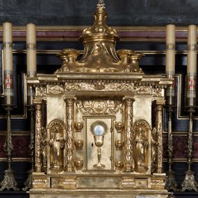 Zdjęcie nr 1: Tabernakulum w formie sześcianu, na niskim cokole, z dwukolumnowym portykiem utworzonym z pilastrów toskańskich i kolumn kompozytowych od frontu i dwóch analogicznych kolumn na tle pilastrów po bokach podtrzymujących przełamujące się na nimi belkowanie. Kolumny osadzone na niskich piedestałach, lekko wybrzuszonych, otoczonych liśćmi akantu. Kolumny pokryte symetrycznie układającymi i przeplatającymi się wokół pionowego trzpienia liśćmi i zwieńczone jońskimi kapitelami. Fryz belkowania zdobiony uskrzydloną główką aniołka, liściastymi wolutami, rautami w drobnych kartuszach i kostkowaniem. Drzwi tabernakulum zdobione są płaskorzeźbionym kielichem z hostią. Po bokach wypukło rzeźbione główki anielskie, a nad nimi okrągły medalion z literami IHS obwiedziony wieńcem laurowym, z elementami przechodzących między sobą listw kartuszowych i podwieszonych pod nie festonów. W ściankach naroży tabernakulum cztery nisze konchowe z muszlami wypełnione figurkami świętych. Jeden w wielbłądziej skórze trzymający księgę z barankiem przedstawia św. Jana Chrzciciela. Trzej pozostali są trudni do identyfikacji. Jeden trzyma otwartą księgę, a u jego stóp widnieje mitra książęca, drugi trzyma pastorał a u jego stóp leży infuła, trzeci z laską i kapeluszem u stóp (św. Jakub?). 


