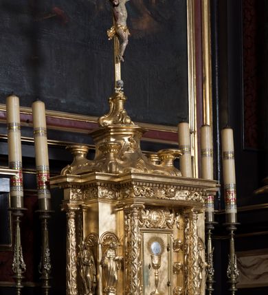 Zdjęcie nr 2: Tabernakulum w formie sześcianu, na niskim cokole, z dwukolumnowym portykiem utworzonym z pilastrów toskańskich i kolumn kompozytowych od frontu i dwóch analogicznych kolumn na tle pilastrów po bokach podtrzymujących przełamujące się na nimi belkowanie. Kolumny osadzone na niskich piedestałach, lekko wybrzuszonych, otoczonych liśćmi akantu. Kolumny pokryte symetrycznie układającymi i przeplatającymi się wokół pionowego trzpienia liśćmi i zwieńczone jońskimi kapitelami. Fryz belkowania zdobiony uskrzydloną główką aniołka, liściastymi wolutami, rautami w drobnych kartuszach i kostkowaniem. Drzwi tabernakulum zdobione są płaskorzeźbionym kielichem z hostią. Po bokach wypukło rzeźbione główki anielskie, a nad nimi okrągły medalion z literami IHS obwiedziony wieńcem laurowym, z elementami przechodzących między sobą listw kartuszowych i podwieszonych pod nie festonów. W ściankach naroży tabernakulum cztery nisze konchowe z muszlami wypełnione figurkami świętych. Jeden w wielbłądziej skórze trzymający księgę z barankiem przedstawia św. Jana Chrzciciela. Trzej pozostali są trudni do identyfikacji. Jeden trzyma otwartą księgę, a u jego stóp widnieje mitra książęca, drugi trzyma pastorał a u jego stóp leży infuła, trzeci z laską i kapeluszem u stóp (św. Jakub?). 


