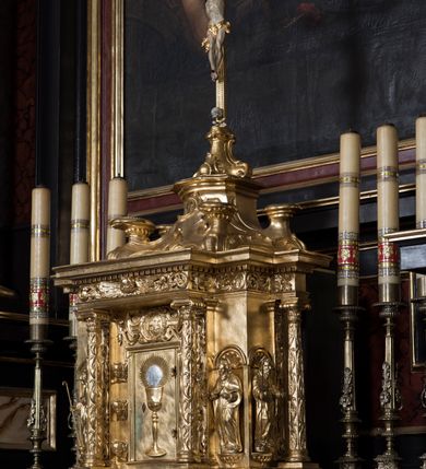 Zdjęcie nr 3: Tabernakulum w formie sześcianu, na niskim cokole, z dwukolumnowym portykiem utworzonym z pilastrów toskańskich i kolumn kompozytowych od frontu i dwóch analogicznych kolumn na tle pilastrów po bokach podtrzymujących przełamujące się na nimi belkowanie. Kolumny osadzone na niskich piedestałach, lekko wybrzuszonych, otoczonych liśćmi akantu. Kolumny pokryte symetrycznie układającymi i przeplatającymi się wokół pionowego trzpienia liśćmi i zwieńczone jońskimi kapitelami. Fryz belkowania zdobiony uskrzydloną główką aniołka, liściastymi wolutami, rautami w drobnych kartuszach i kostkowaniem. Drzwi tabernakulum zdobione są płaskorzeźbionym kielichem z hostią. Po bokach wypukło rzeźbione główki anielskie, a nad nimi okrągły medalion z literami IHS obwiedziony wieńcem laurowym, z elementami przechodzących między sobą listw kartuszowych i podwieszonych pod nie festonów. W ściankach naroży tabernakulum cztery nisze konchowe z muszlami wypełnione figurkami świętych. Jeden w wielbłądziej skórze trzymający księgę z barankiem przedstawia św. Jana Chrzciciela. Trzej pozostali są trudni do identyfikacji. Jeden trzyma otwartą księgę, a u jego stóp widnieje mitra książęca, drugi trzyma pastorał a u jego stóp leży infuła, trzeci z laską i kapeluszem u stóp (św. Jakub?). 


