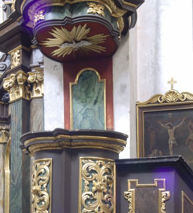 Zdjęcie nr 2: Ambona wisząca, przyścienna, umieszczona na ścianie łuku tęczowego, z parapetem dziś nieistniejących schodów od strony prezbiterium i usytuowanej przy nim kaplicy. Kosz ambony założony na rzucie trójliścia z trójkątnymi występami, wsparty na puklowanej konsoli z podwieszoną szyszką. Dolna część ambony wybrzuszona, pokryta rozłożystymi liśćmi akantu, kosz prosty na rzucie odcinków koła z prostokątnymi płycinami wypełnionymi liśćmi akantu, zakończony profilowanym gzymsem. Zaplecek z profilowaną płyciną zakończoną łukiem półkolistym nadwieszonym. Nad koszem baldachim powielający rzut kosza, zwieńczony silnie wystającym gzymsem, z podwieszoną w podniebieniu gołębicą Ducha Świętego na tle glorii promienistej. Nad baldachimem kabłąki w formie spływów wolutowych opatrzonych liśćmi akantu, podtrzymujące koronę zamkniętą z kulą i krzyżem. Parapet schodów z dekoracją w postaci trzech płycin w kształcie równoległoboku obwiedzionych profilowanymi ramami. Płycina w polu łączącym parapet schodów z podestem przed amboną w formie stojącego prostokąta z uszakami, ujętego motywami roślinnymi. Ambona w kolorze czarnym, z zielonymi i czerwonymi imitacjami marmoryzacji, ze złoconymi ornamentami. 